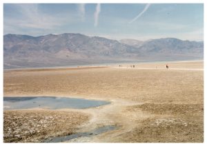 photo of badwater basin death valley national park with people walking across the saltpan to the lake surrounded by mountains and contrails in the sky
