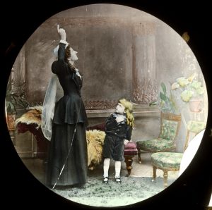 In drawing room setting, in 1900, middle-aged woman in long back skirt and jacket, with long white veil, points to the ceiling, watched by young boy, with long hair in ringlets, and dressed in dark green velvet knickerbocker suit; right hand over his heart, left hand in his trouser pocket.