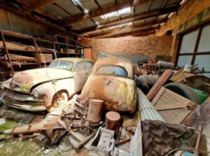 Disused and dilapidated shed, with two dusty, rusty car bodies dating from 1950s and 1960s, and various building offcuts and detritus.