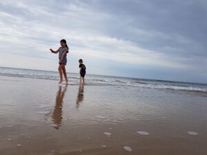 Two children - one a girl of about 8 and the other a younger boy - holding their clothes around their knees face a pristine empty beach as a shallow wave recedes from a sandy expanse of beach.